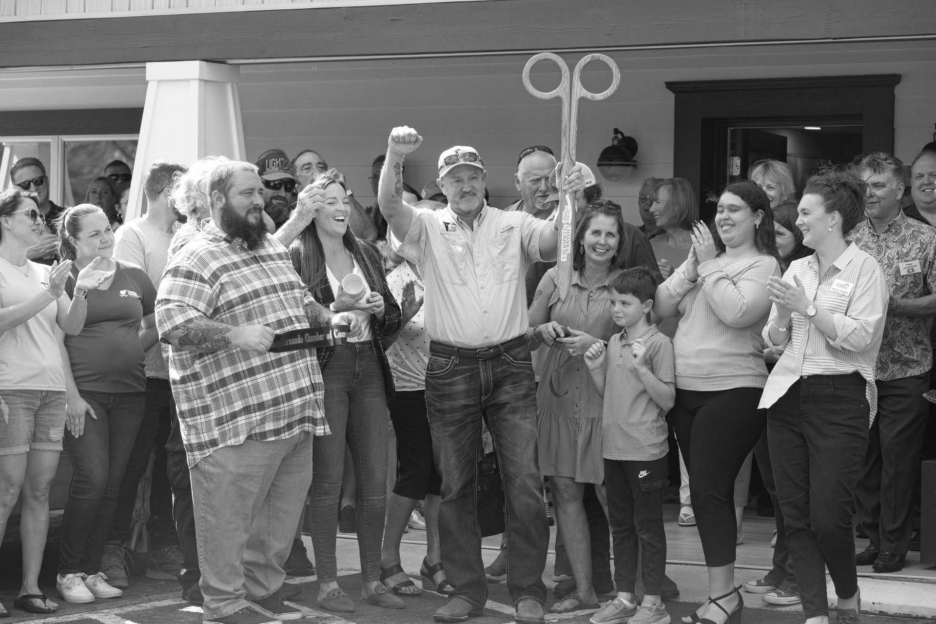People at a ribbon-cutting ceremony. Man with large scissors raises arm. Crowd applauds in front of a building.