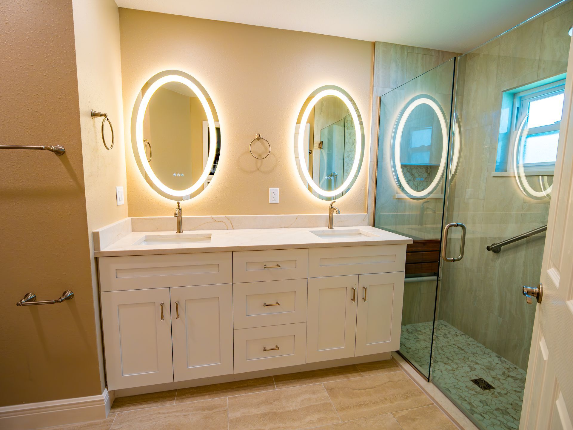 Bathroom with white vanity, oval lit mirrors, glass shower, and tan tile.