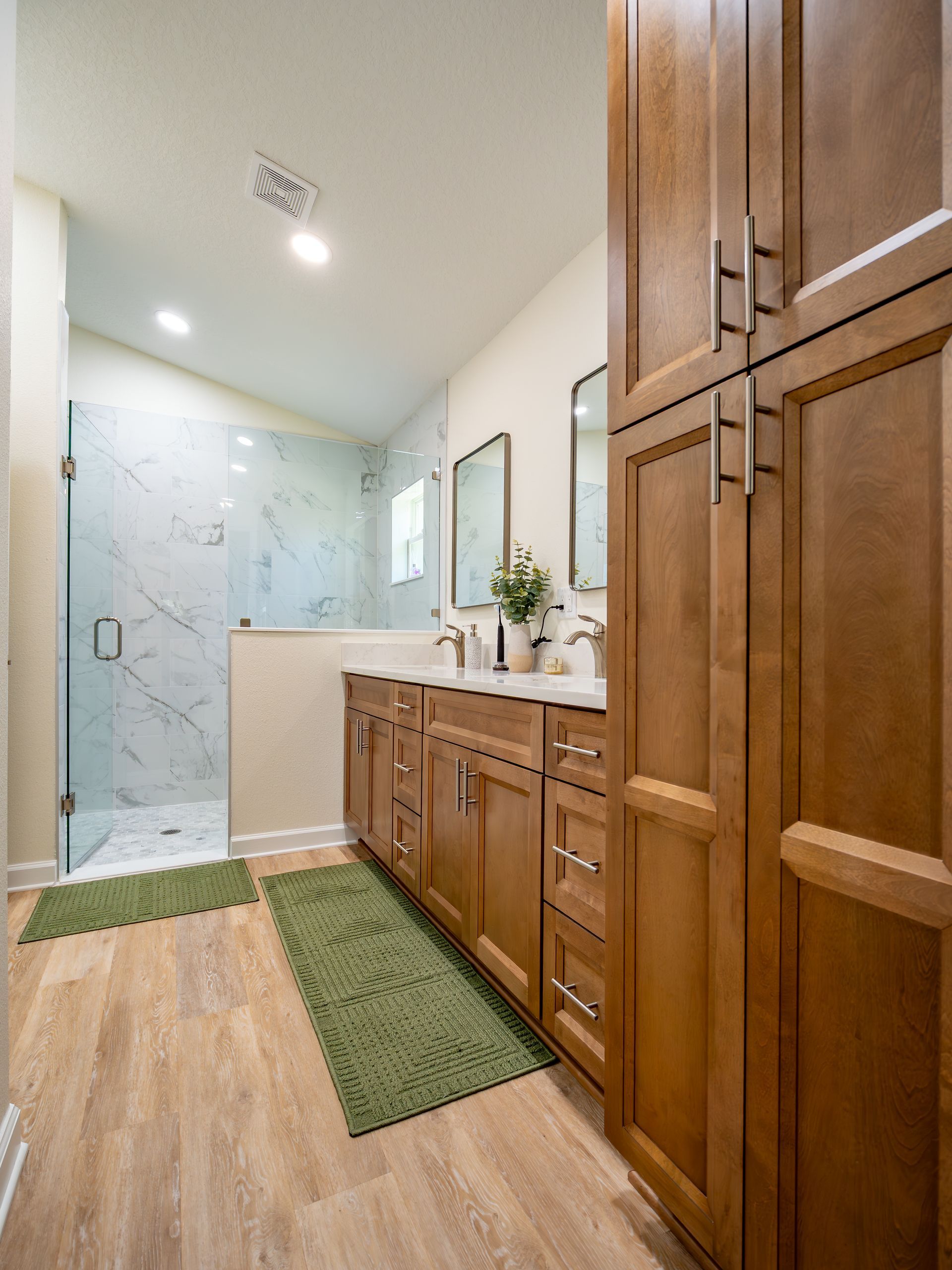 Bathroom with wooden cabinets, marble shower, and wood-look flooring. Green bath mats add color.