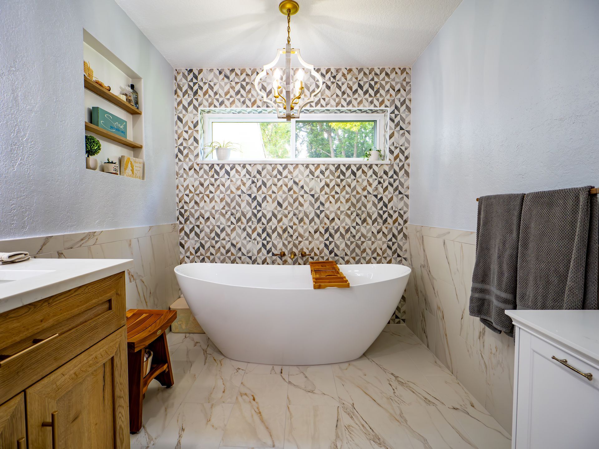 Bathroom with a white tub, marble-look tile, patterned accent wall, wooden vanity, and gold chandelier.