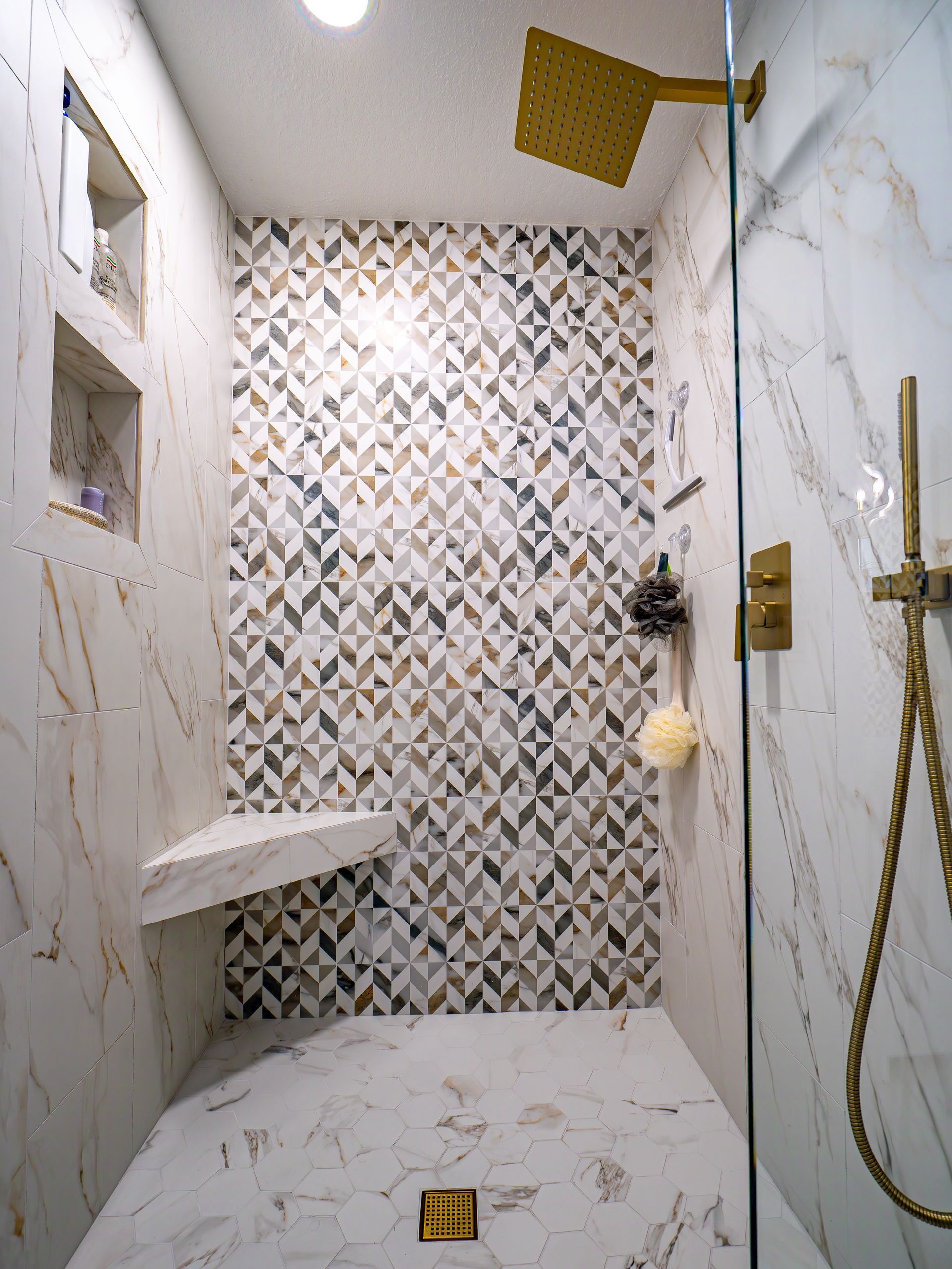 Shower with marble tile, gold fixtures, geometric accent wall, corner seat.