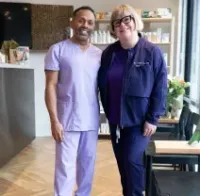Two people standing together in a bright café, wearing purple outfits and smiling at the camera.