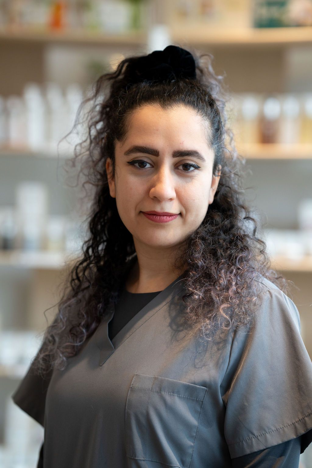 Person with curly hair in gray scrubs, standing indoors with shelves softly blurred in the background.
