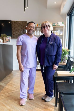 Two people standing together in a bright salon, one in lavender scrubs and one in a navy jacket.