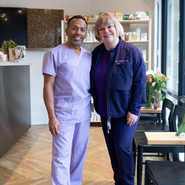 Two healthcare workers in scrubs standing together in a clinic lobby, smiling.