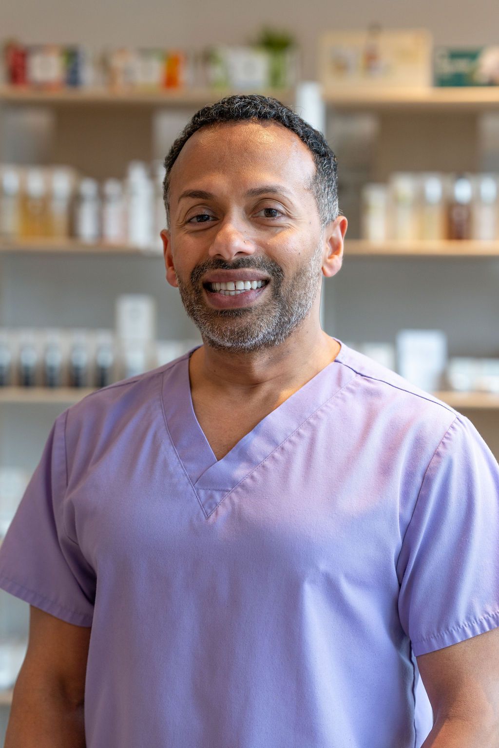 Smiling person in purple scrubs standing in a pharmacy with shelves of medicine behind them