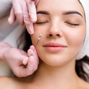 Gloved hands hold a fine needle near a woman’s cheek during a cosmetic injection treatment