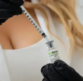 Gloved hands drawing liquid into a syringe from a vial, with a blurred person in the background