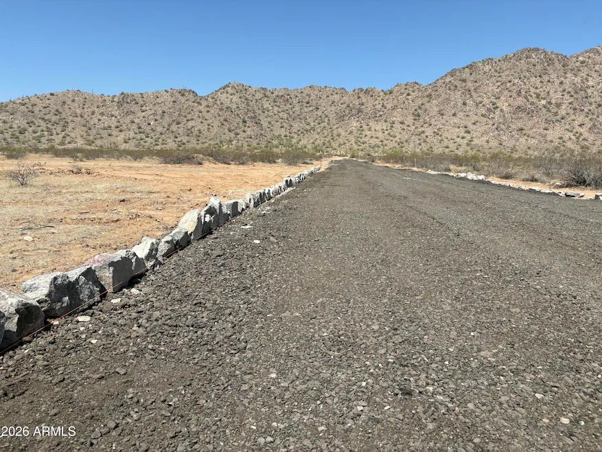 A gravel road borders a desert landscape with rocks lining the edge, leading toward a distant mountain range.