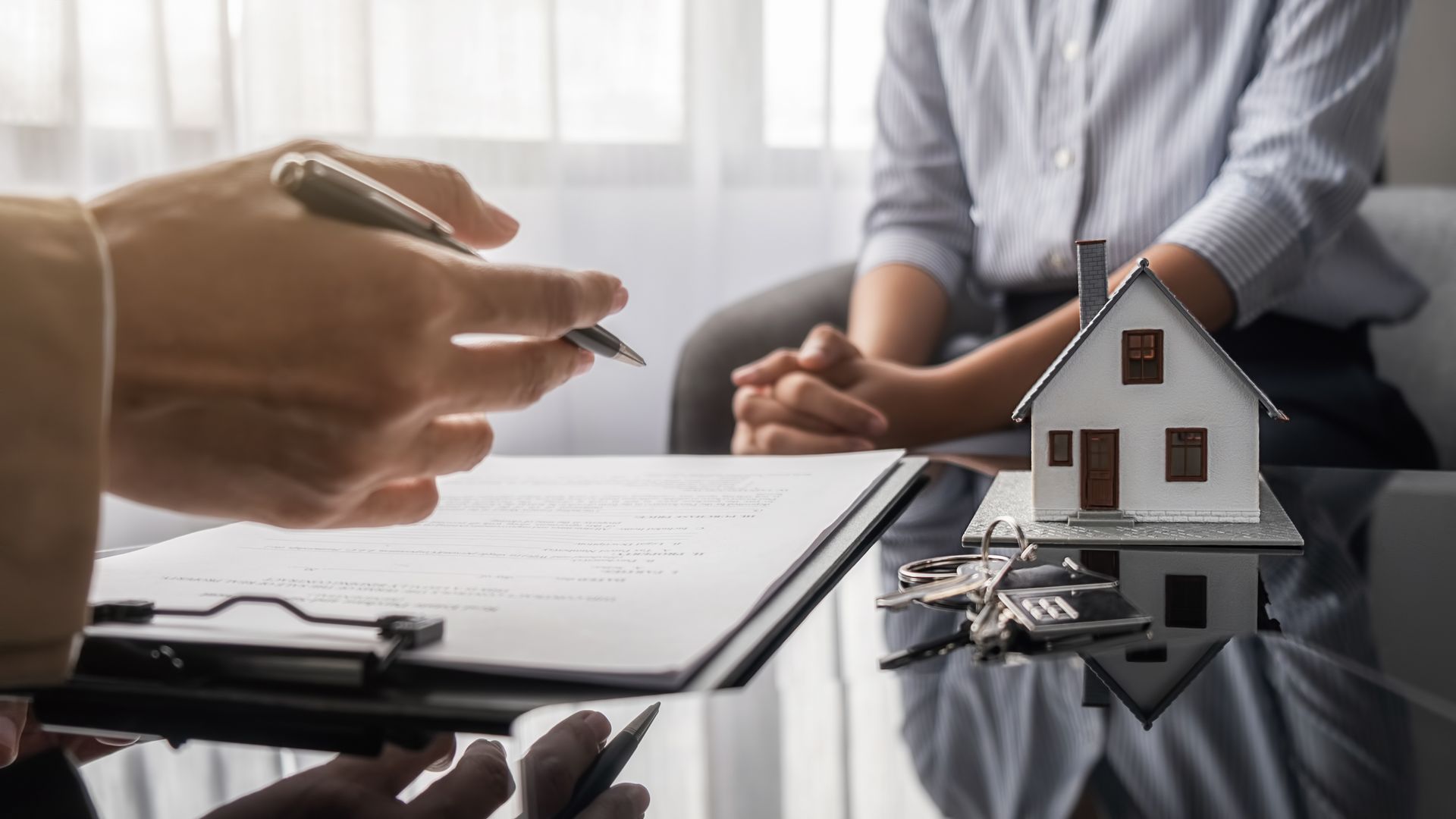 A person holds a pen over a document while sitting across from someone near a model house and keys on a table.