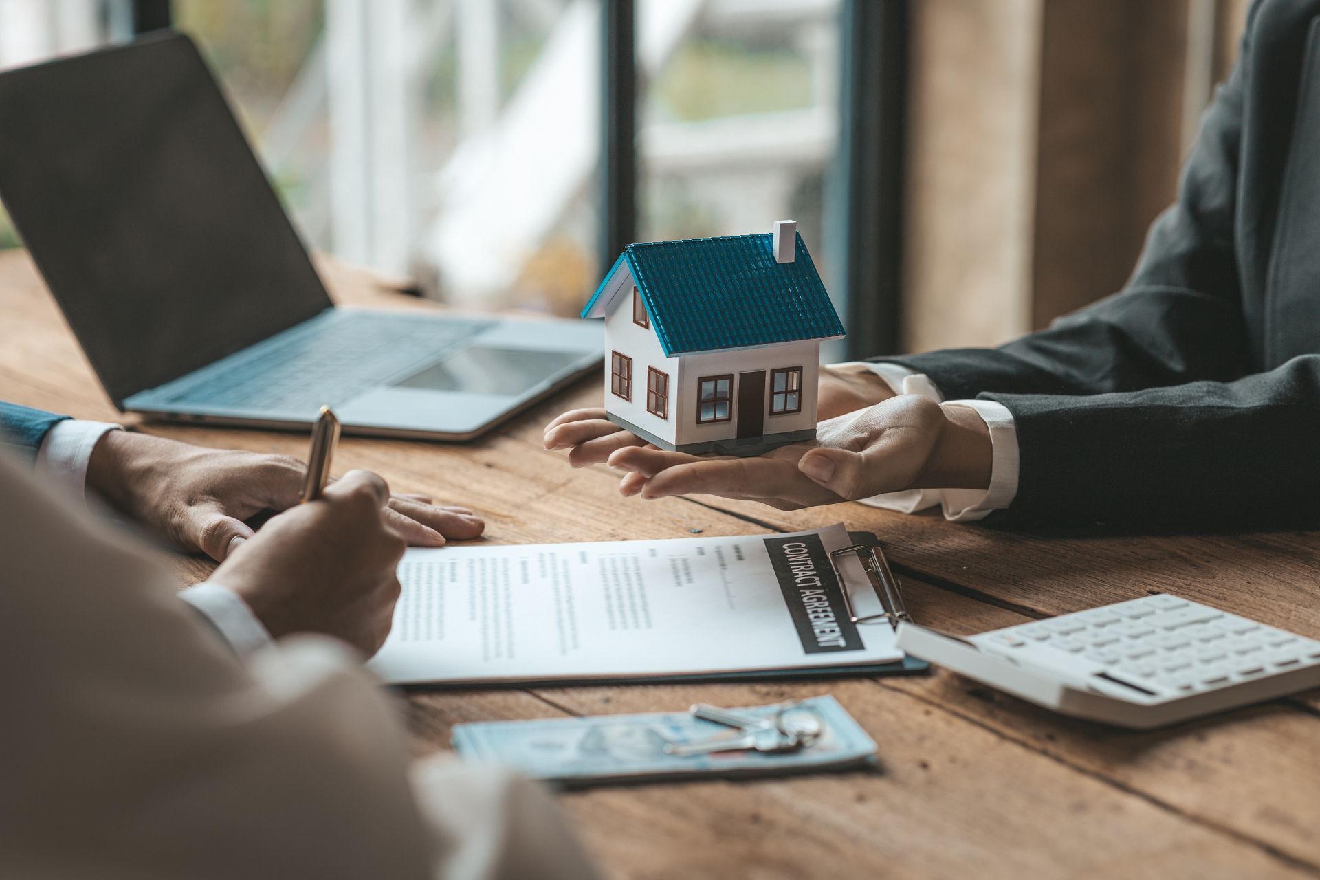 Hands hold a small house model over a contract on a wooden table with a laptop, calculator, and keys nearby.