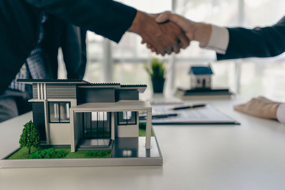 Two people in business suits shake hands over a desk featuring a miniature modern house model and paperwork.