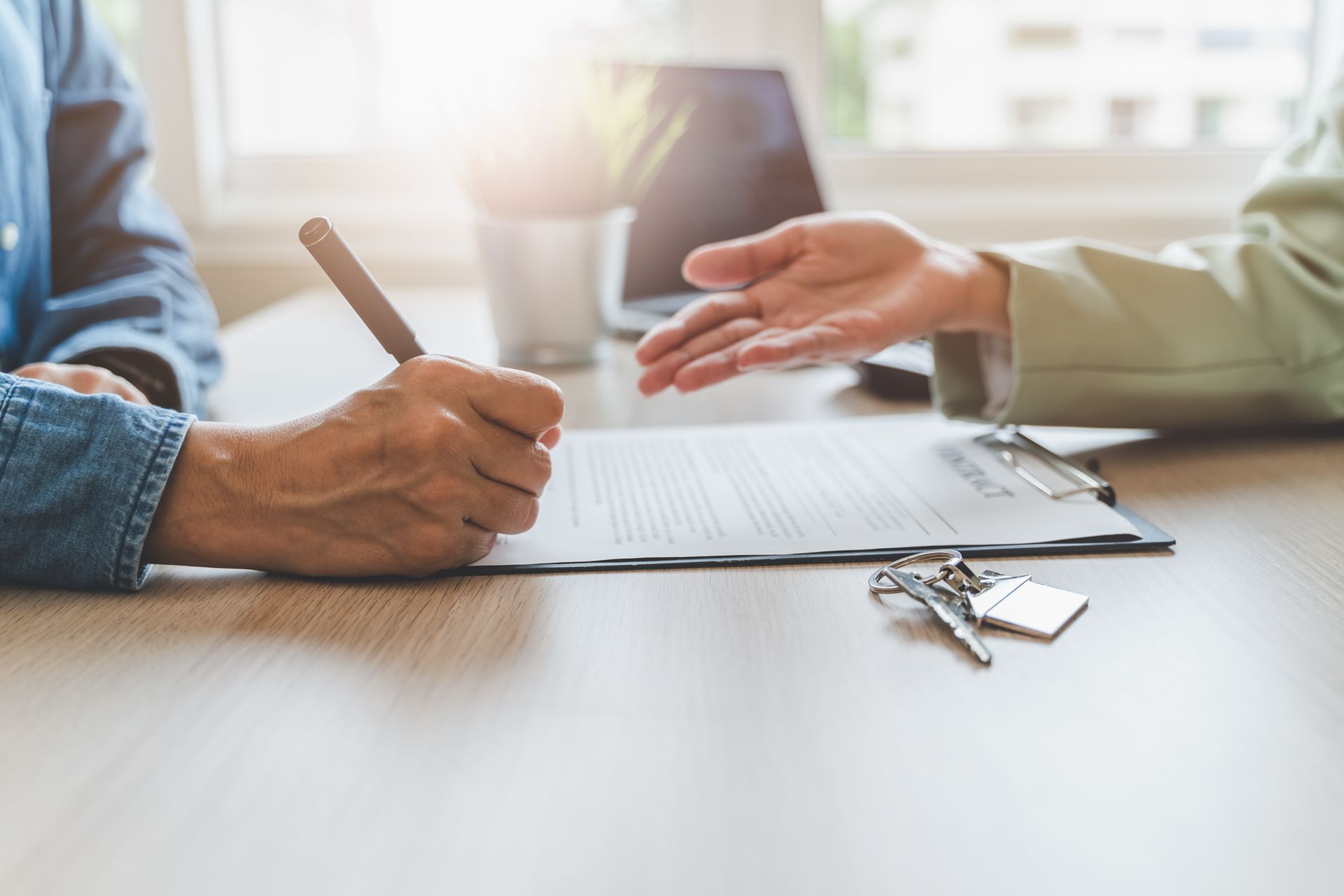 Hands sign a document on a wooden table with keys nearby, while another person gestures toward the paper.
