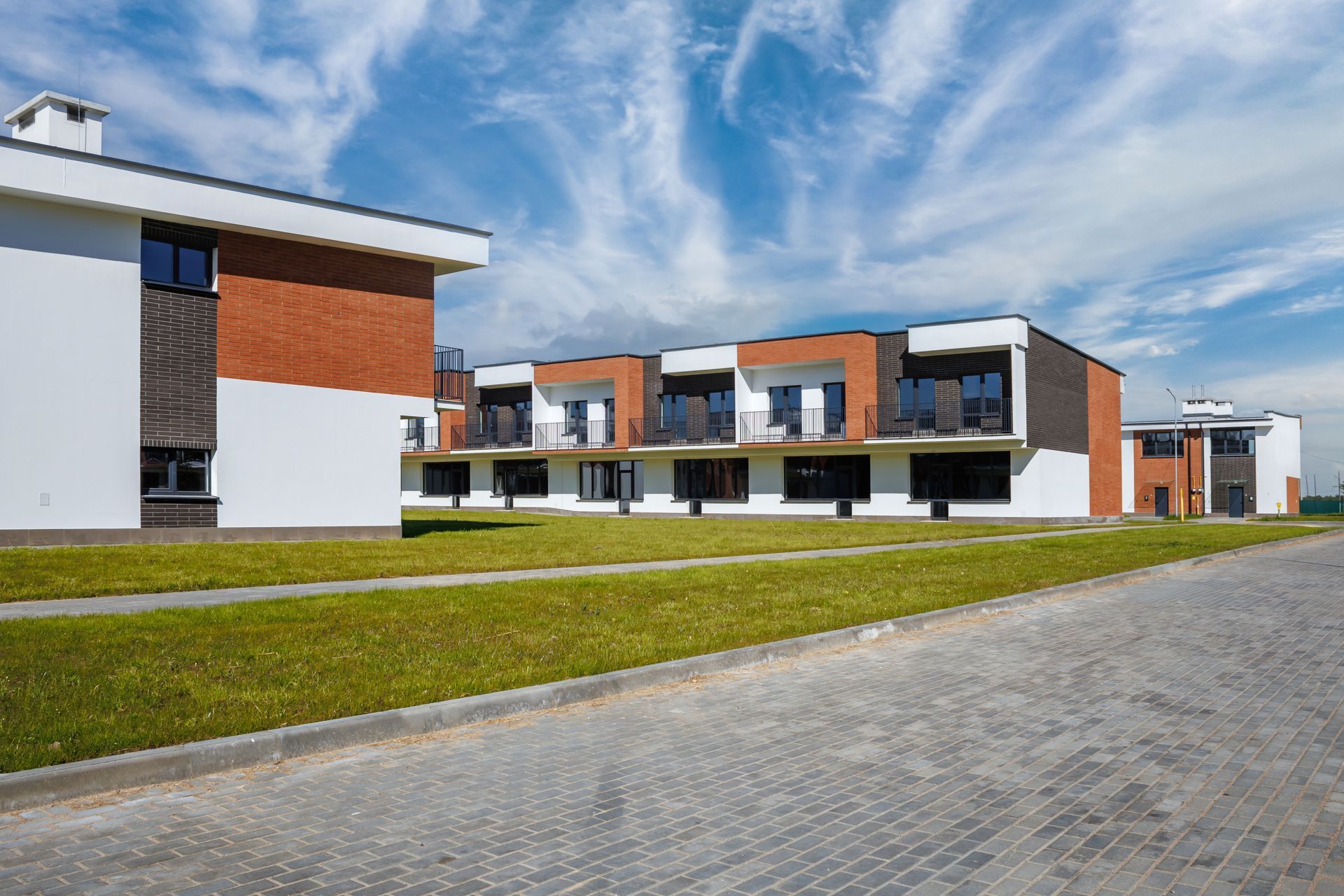 Modern townhouses with brick and white siding, balconies, and large windows set against a blue sky with thin clouds.