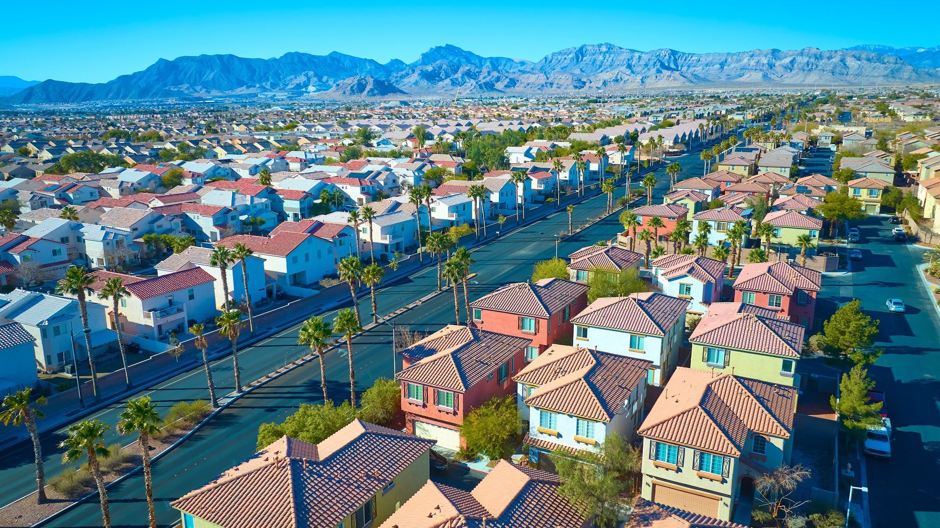 An aerial view of a suburban neighborhood with rows of houses, palm trees, and mountain ranges in the distance.