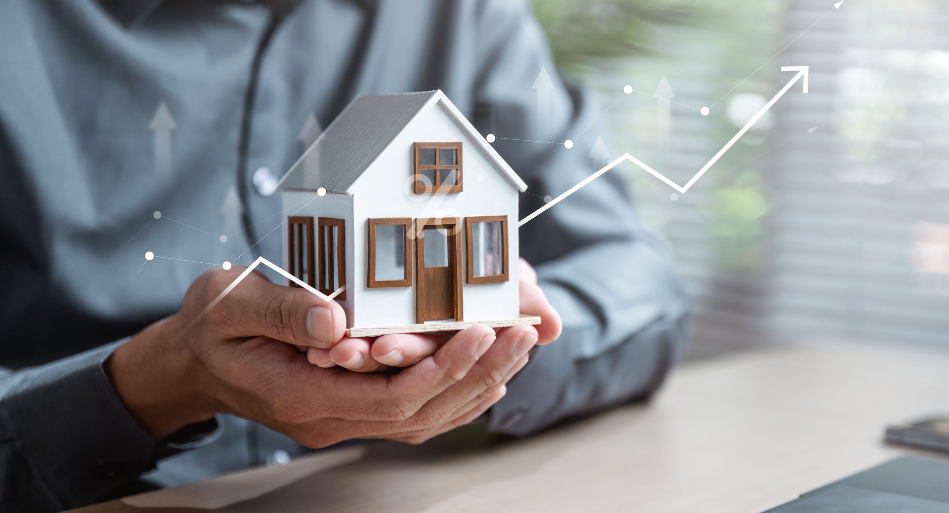 A person holds a small model house in their hands, with a digital graphic of an upward-trending arrow overlaid.