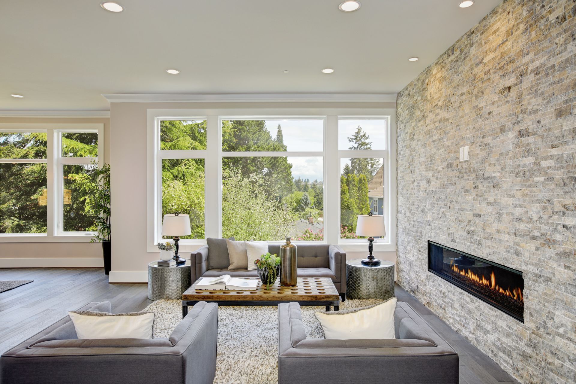 A modern living room with gray furniture, a wooden coffee table, and a linear fireplace set into a textured stone wall.