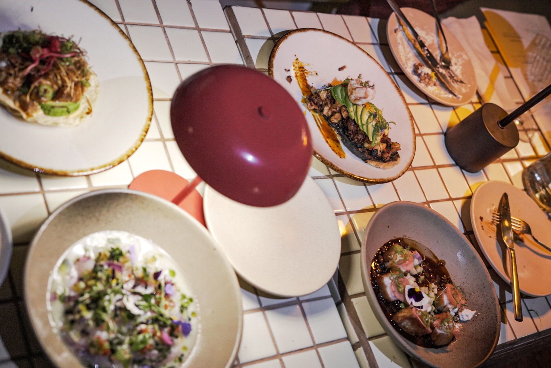 A variety of gourmet dishes are served on white tiled tables, illuminated by a small, round red-topped lamp.