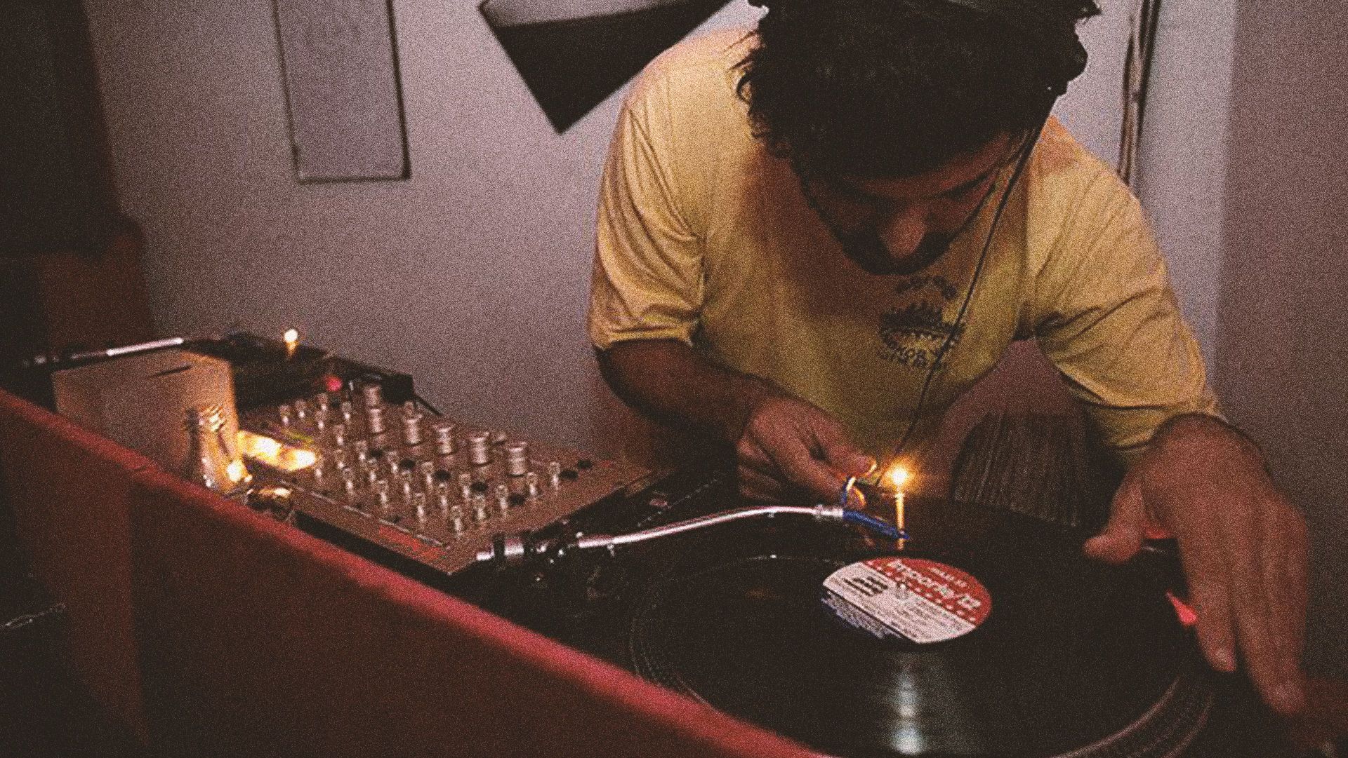 A person in a yellow shirt uses a lighter to illuminate a vinyl record on a turntable in a dimly lit, cozy room.