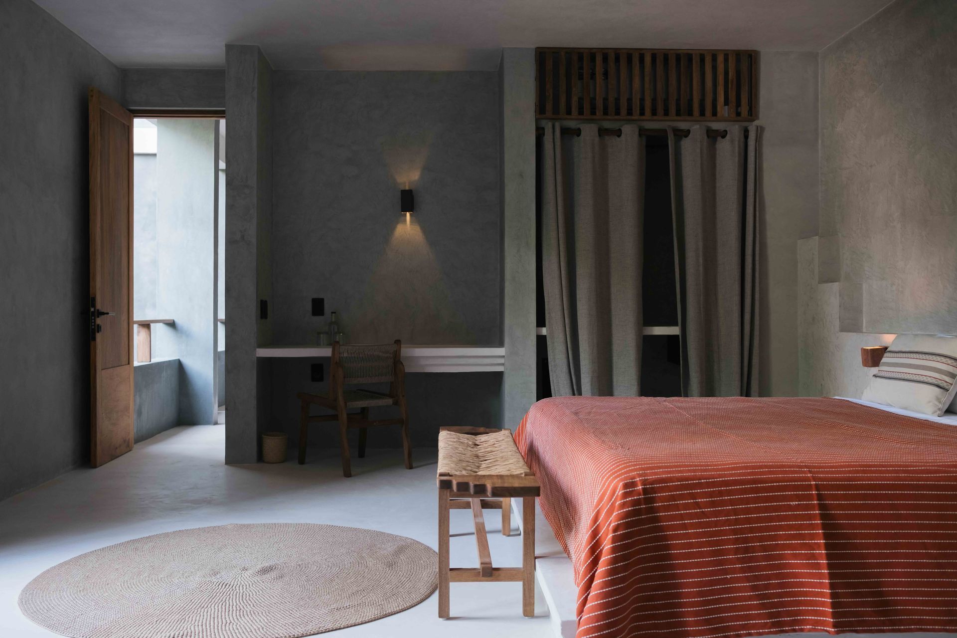 Bedroom with orange-red bedspread, wooden bench, open door to balcony, gray walls, and round rug.