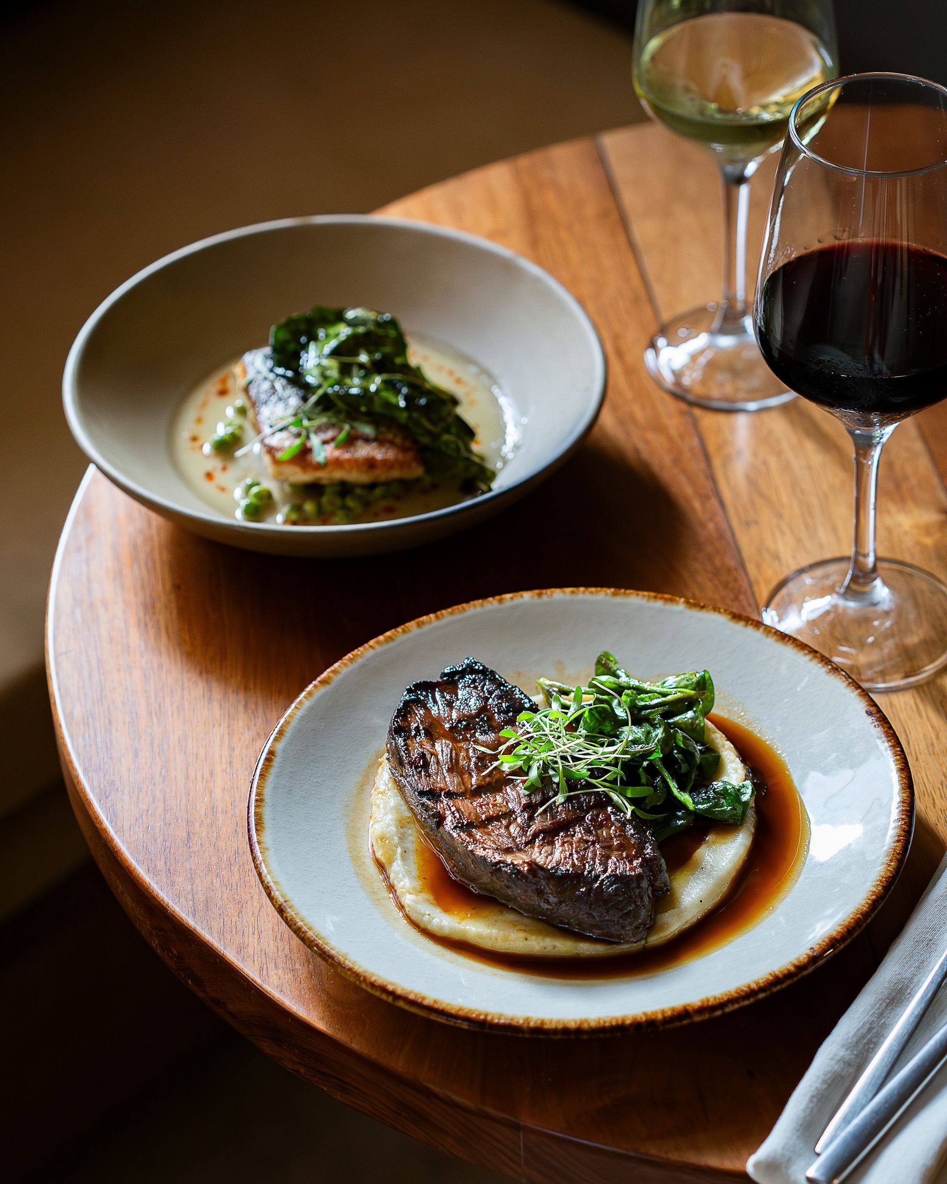 Two plated meals with wine glasses on a wooden table. Steak with sauce and greens, salmon with greens and peas.