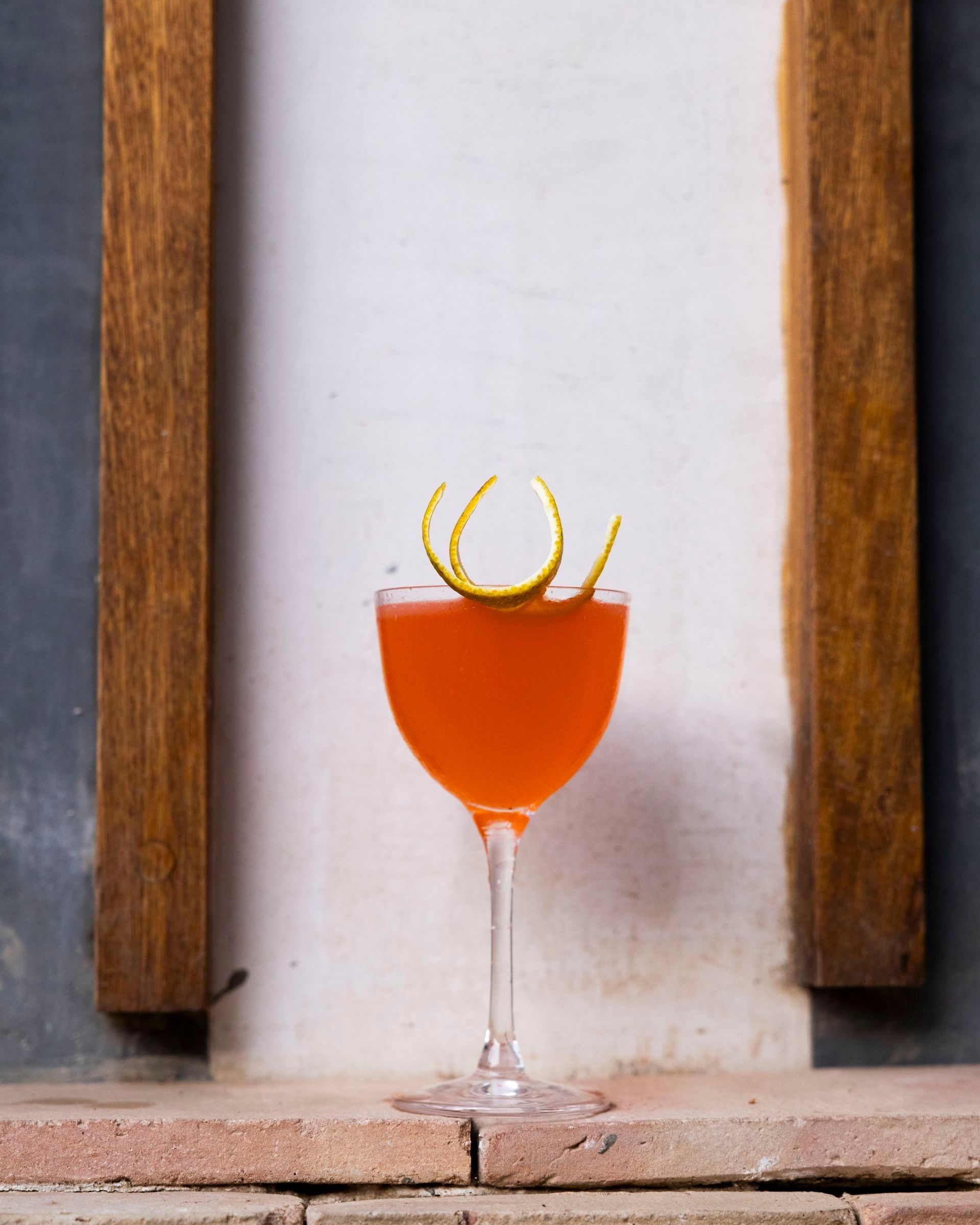 Cocktail in a stemmed glass with orange liquid, garnished with citrus peel, set against a rustic background.
