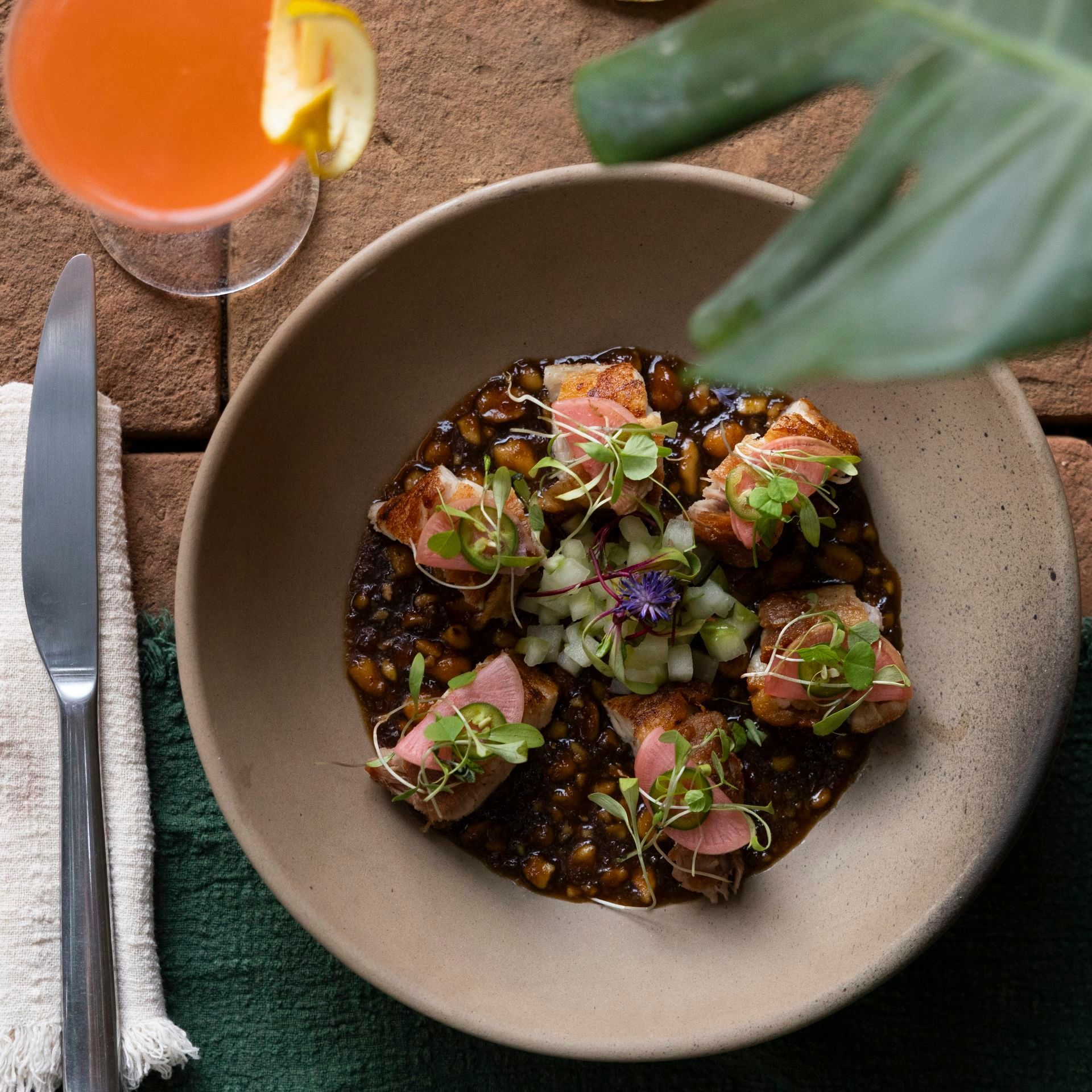 Close-up of a plate of food with pork and lentils, garnished with sprouts, next to a cocktail and a knife.