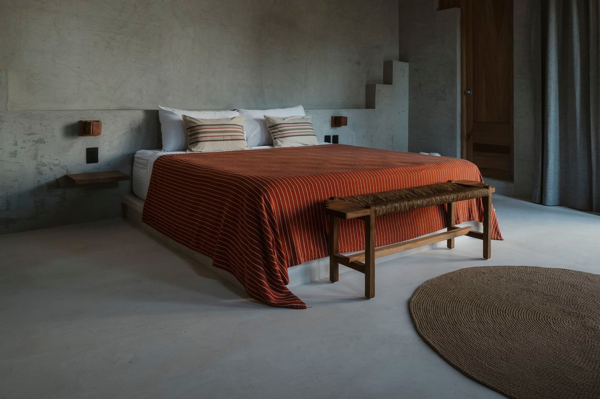 Bedroom with concrete walls, orange patterned bedspread, wooden bench, round rug.