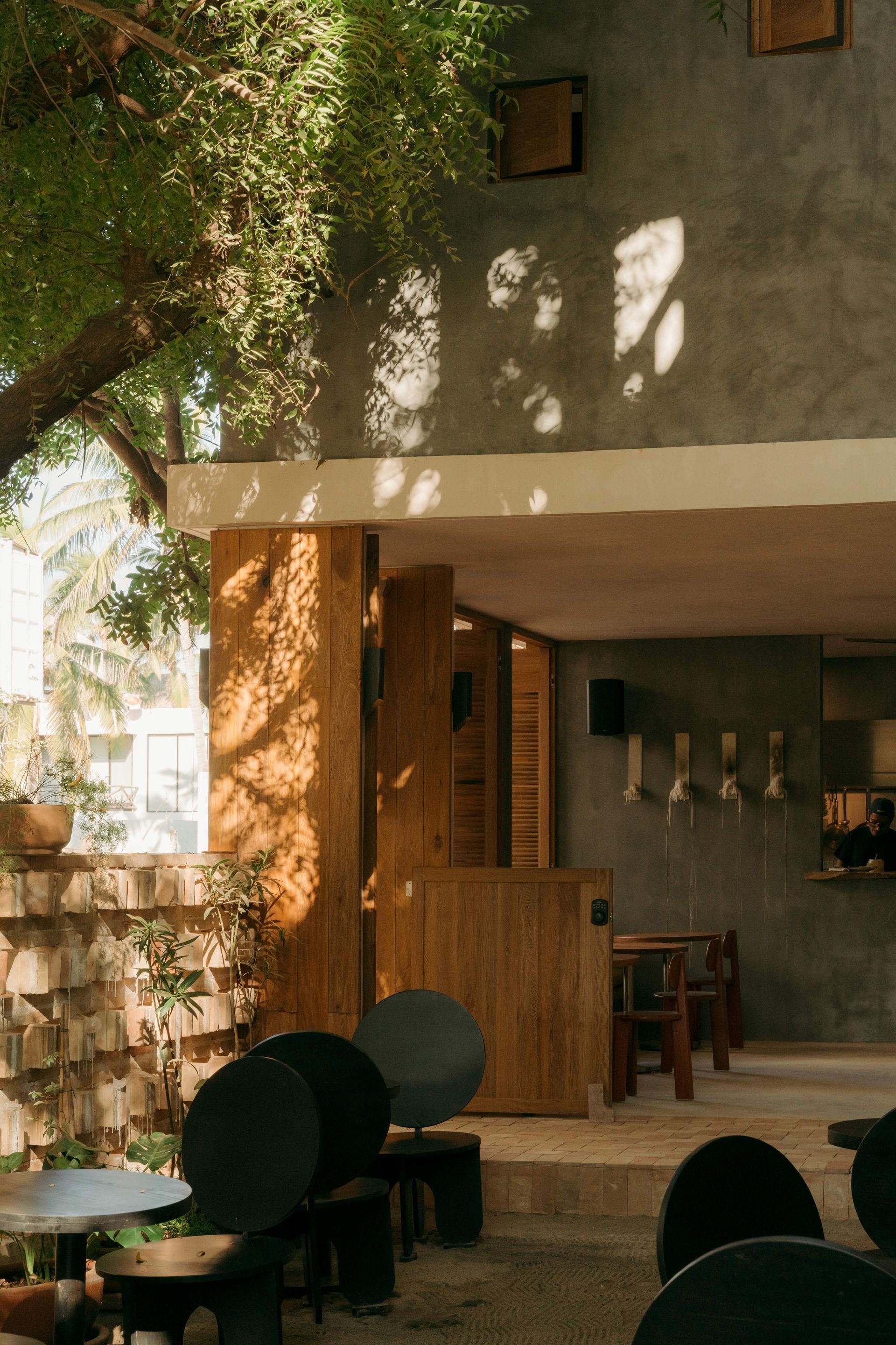 An outdoor cafe patio with wooden accents, gray concrete walls, and dark circular tables and chairs under a leafy tree.