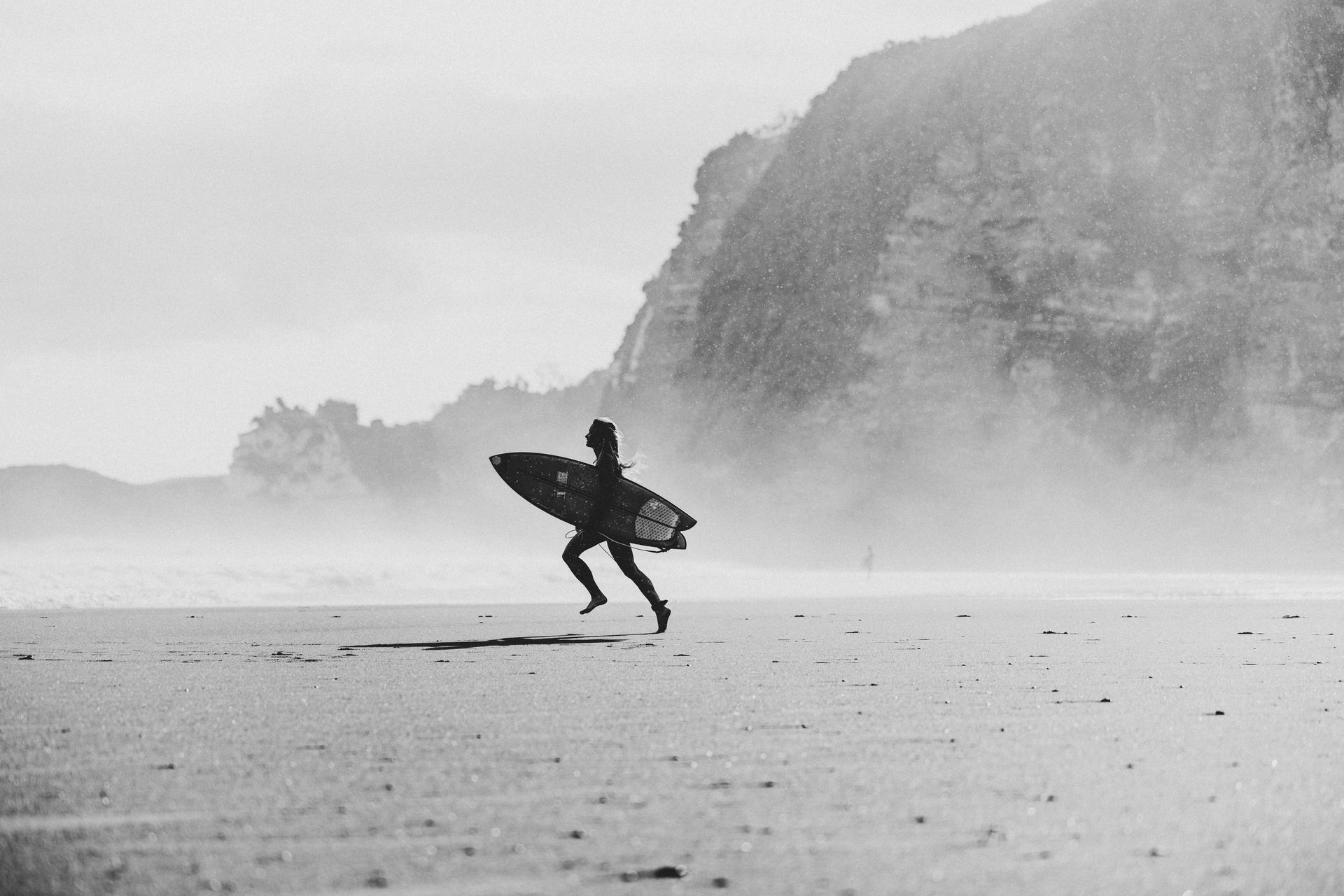A silhouetted person carries a surfboard while running across a foggy beach toward the ocean with a cliff in the background.