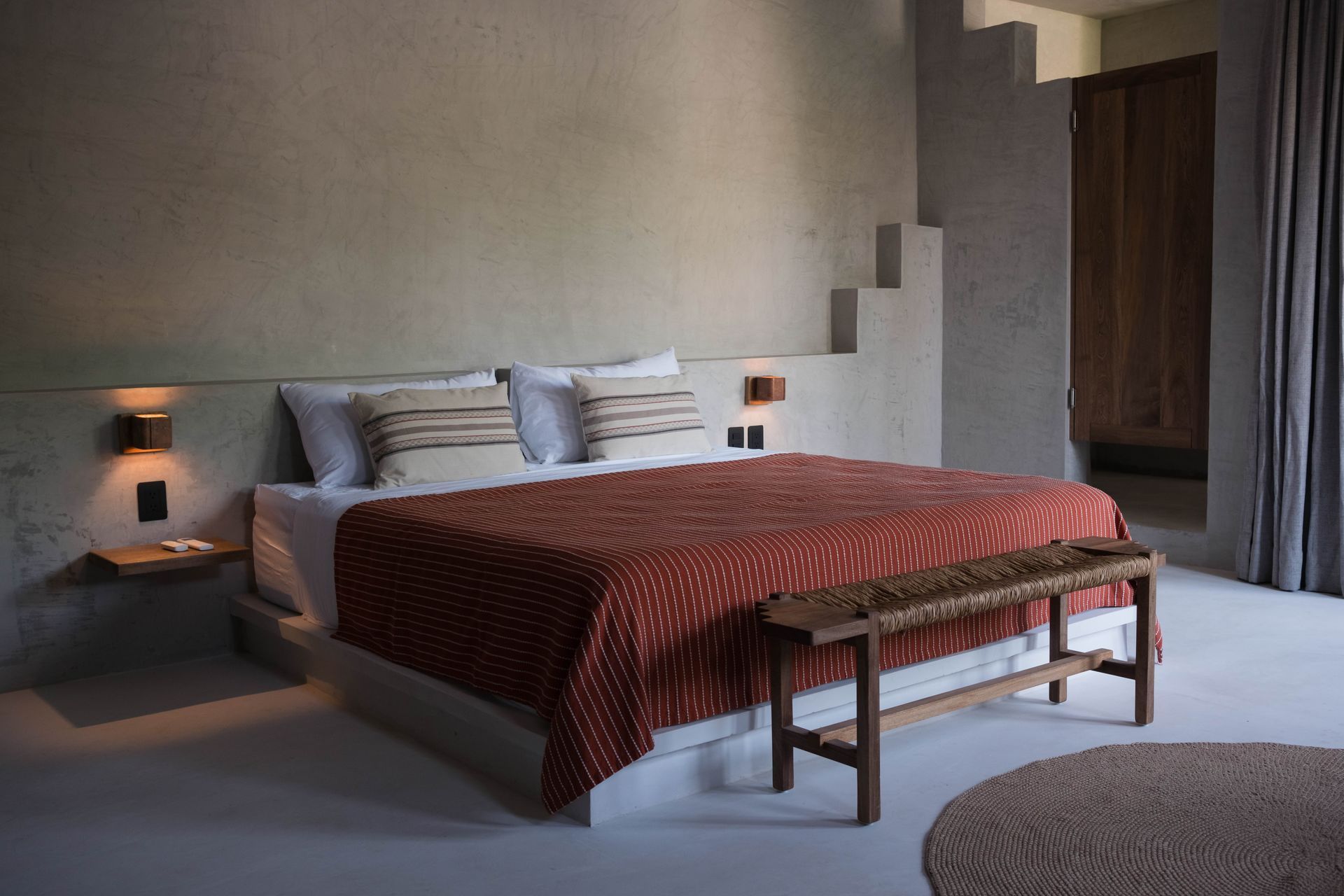 Bedroom with a bed, red quilt, wooden bench, and gray walls. Includes a round rug and side tables.