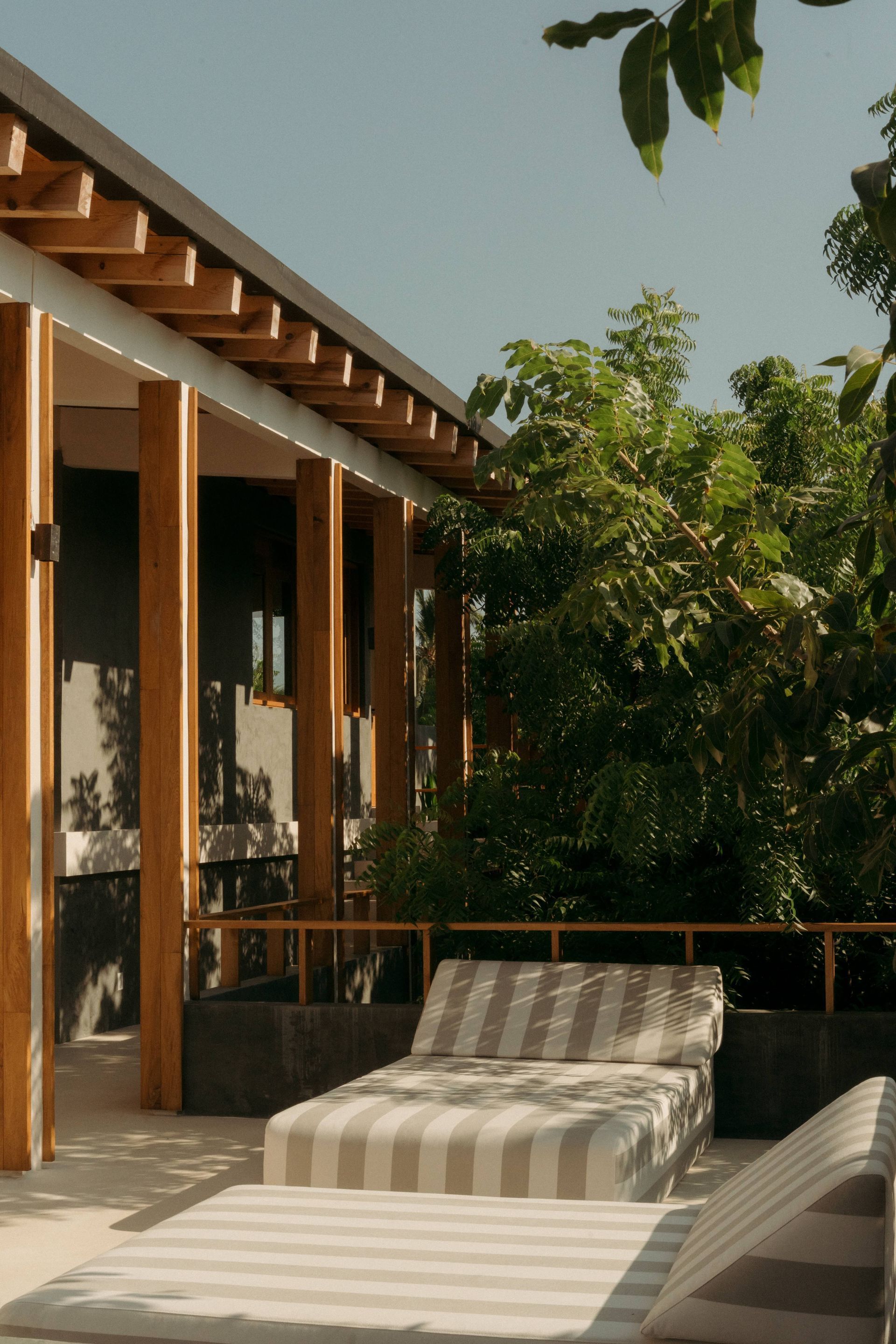 Poolside lounge chairs under a wooden pergola, with lush greenery and a clear sky.