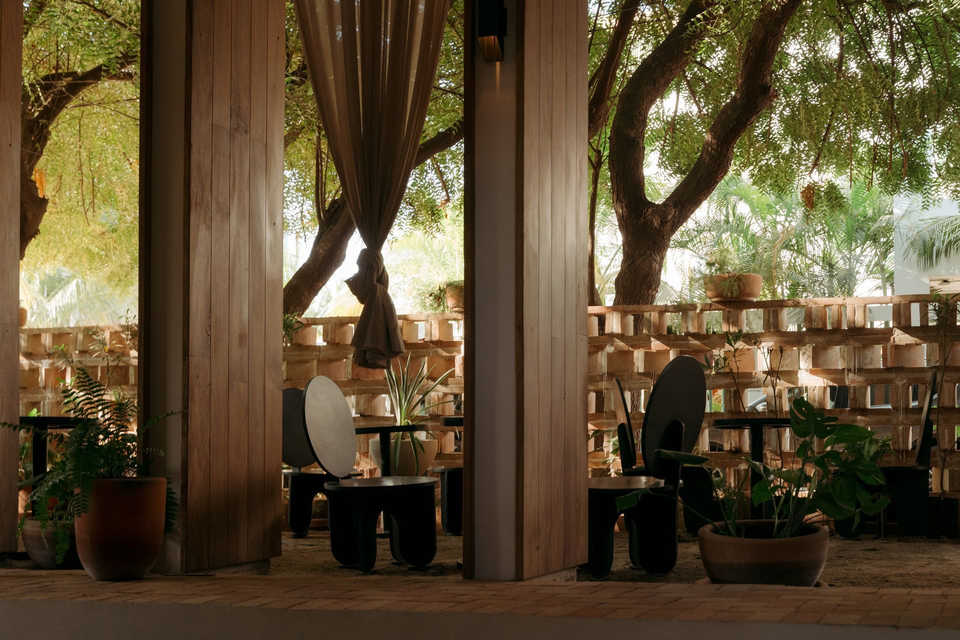 Outdoor dining area, wooden pillars, tables, chairs, trees in background, brick wall, plants, natural light.
