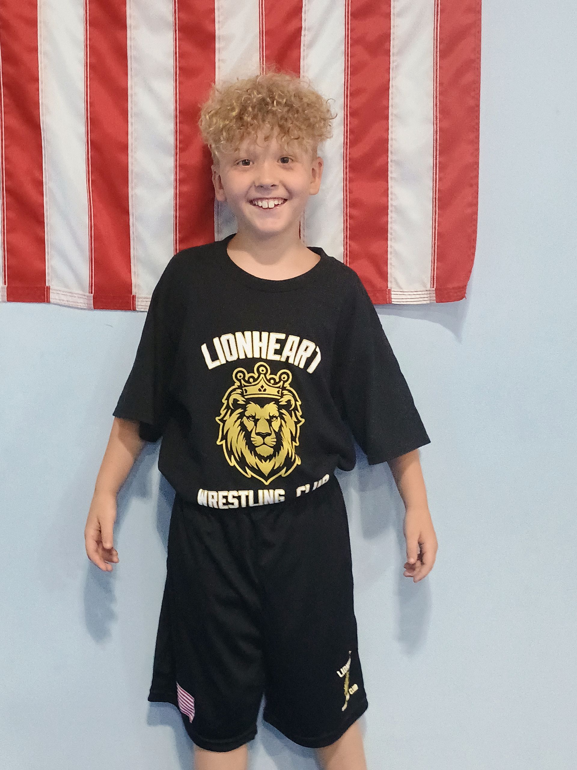 Boy in black wrestling uniform, smiling, standing in front of an American flag.