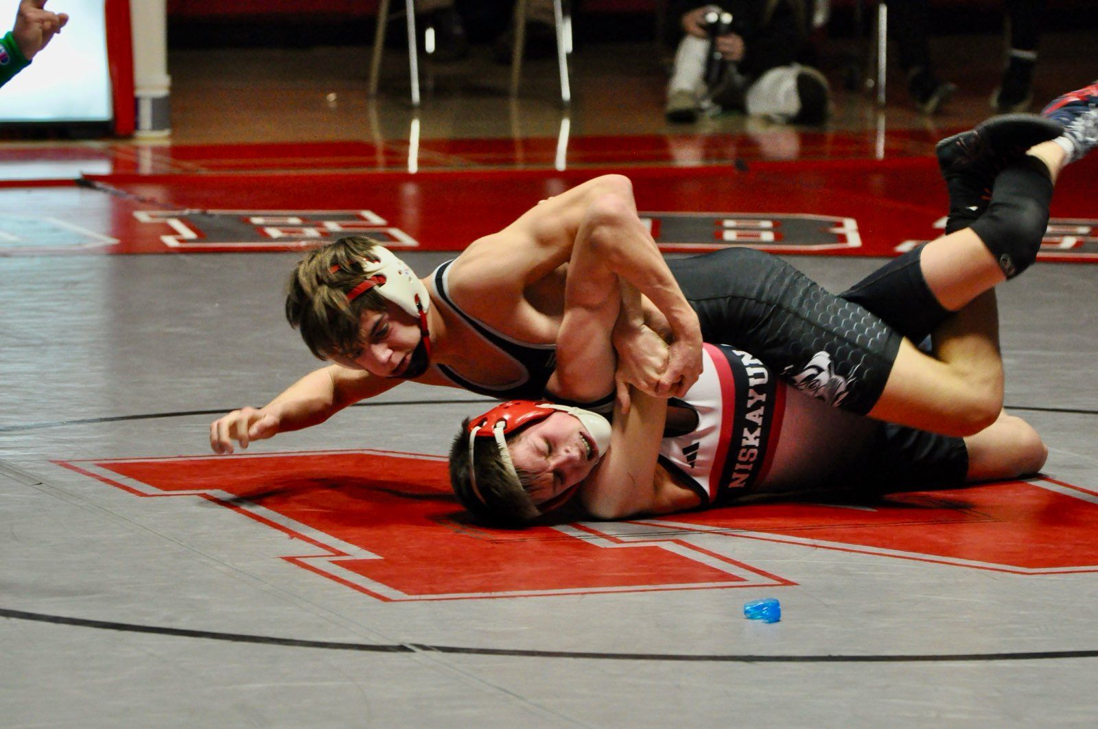 Wrestling coaches instructing a group of children on a wrestling mat in a gym.