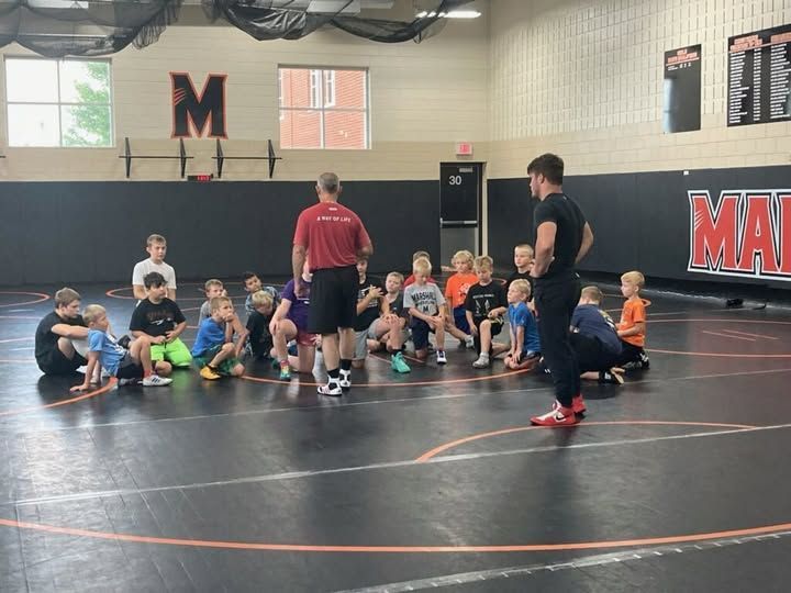 Wrestling coaches instructing a group of children on a wrestling mat in a gym.