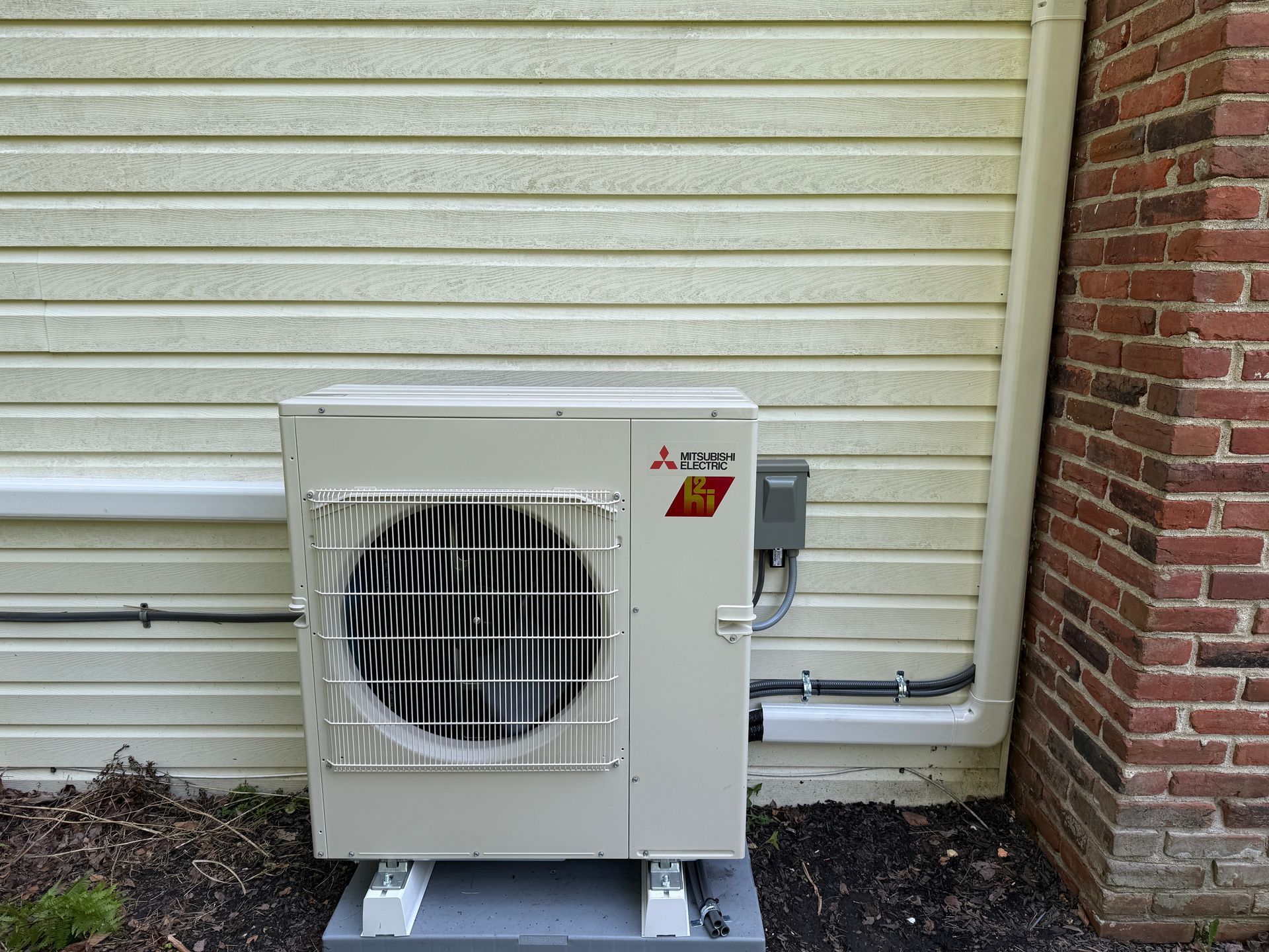 Mitsubishi heat pump unit outside a building with white siding and a brick wall.