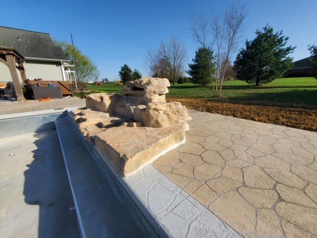 A large rock in the middle of a patio next to a pool