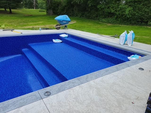 A blue swimming pool with steps and a wheelbarrow in the background.