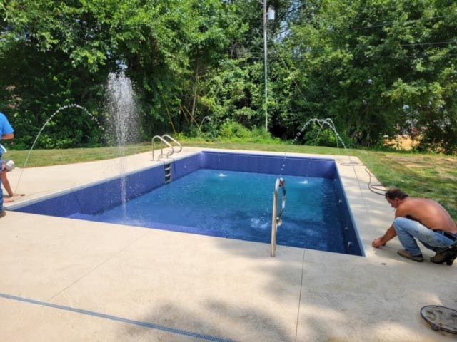 A man is kneeling in front of a swimming pool with a fountain in the background.