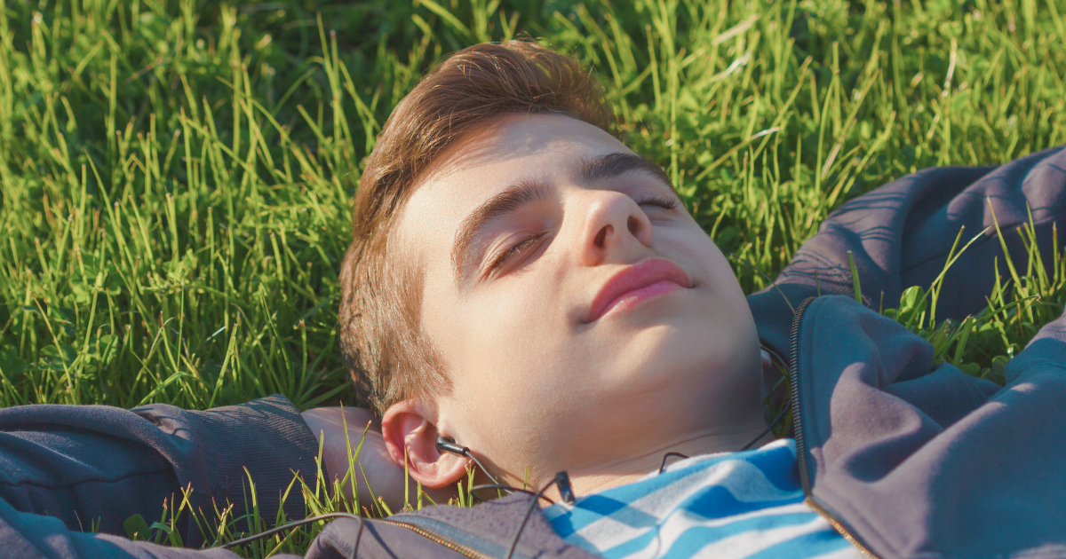 Caucasian teen boy resting in the grass, eyes closed and arms under his head.