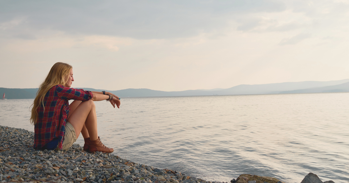 a young blonde woman sitting on the edge of a calm lake