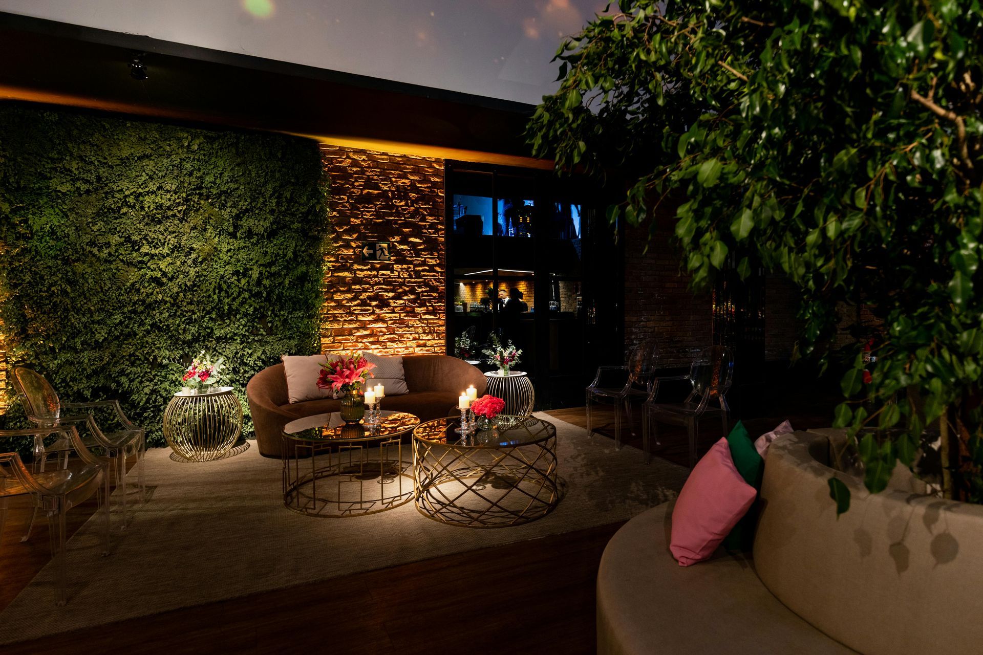 Dim patio lounge with wicker chairs, candles, flowers, and warm accent lighting beside greenery at night