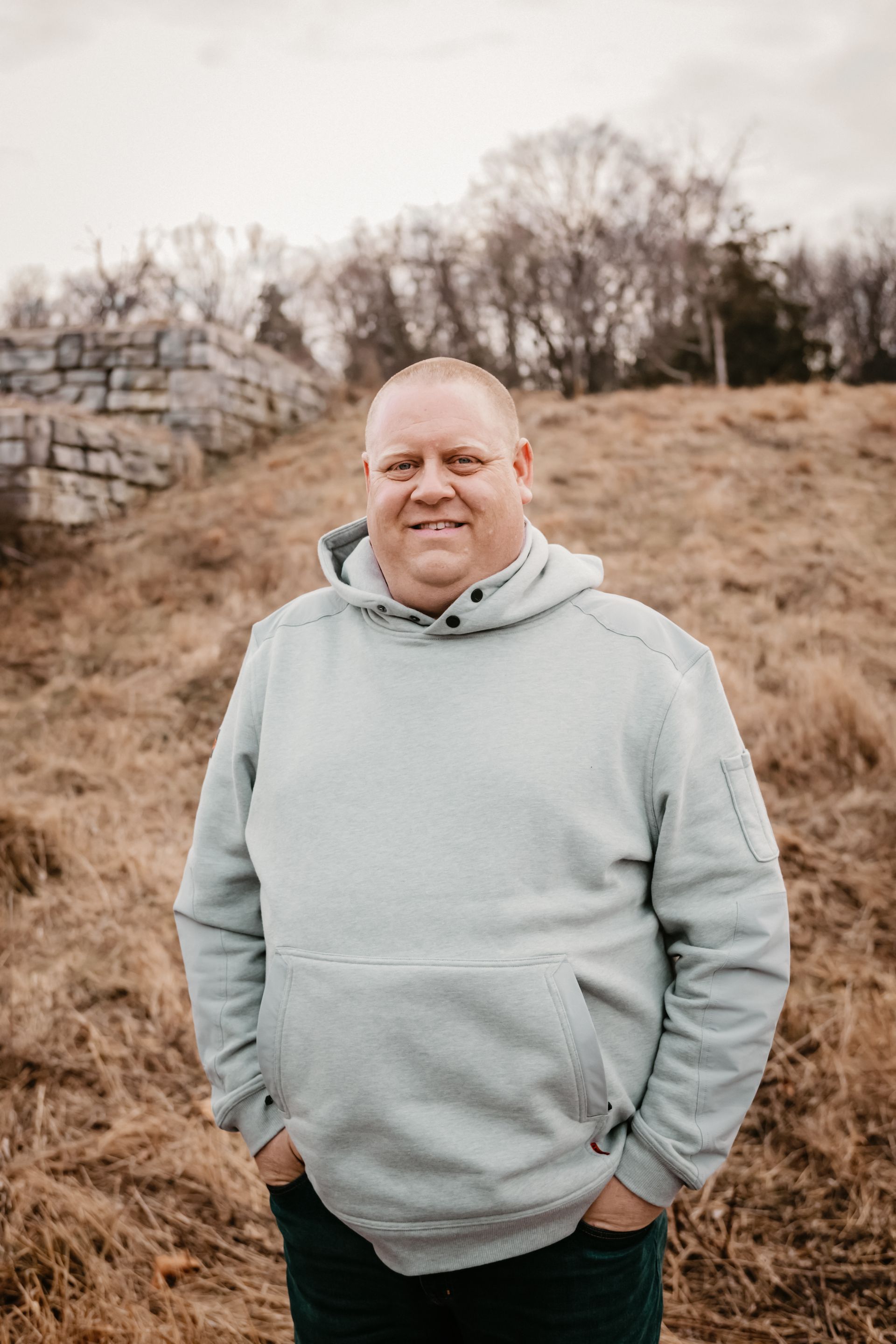 Man in light gray hoodie, standing outdoors, hands in pockets, smiling.