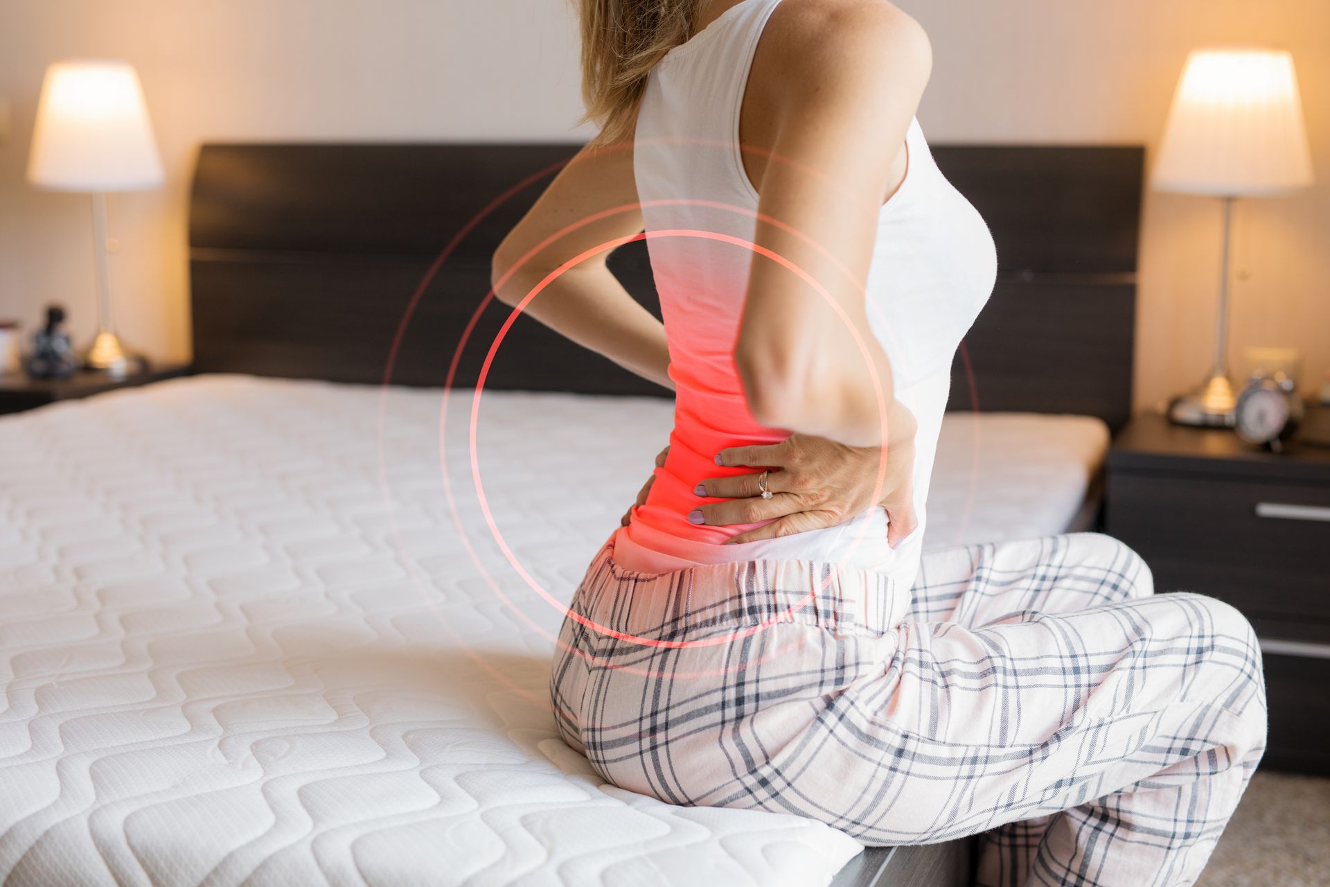 Woman sitting on a bed, holding her lower back in pain; red circles highlight the affected area.