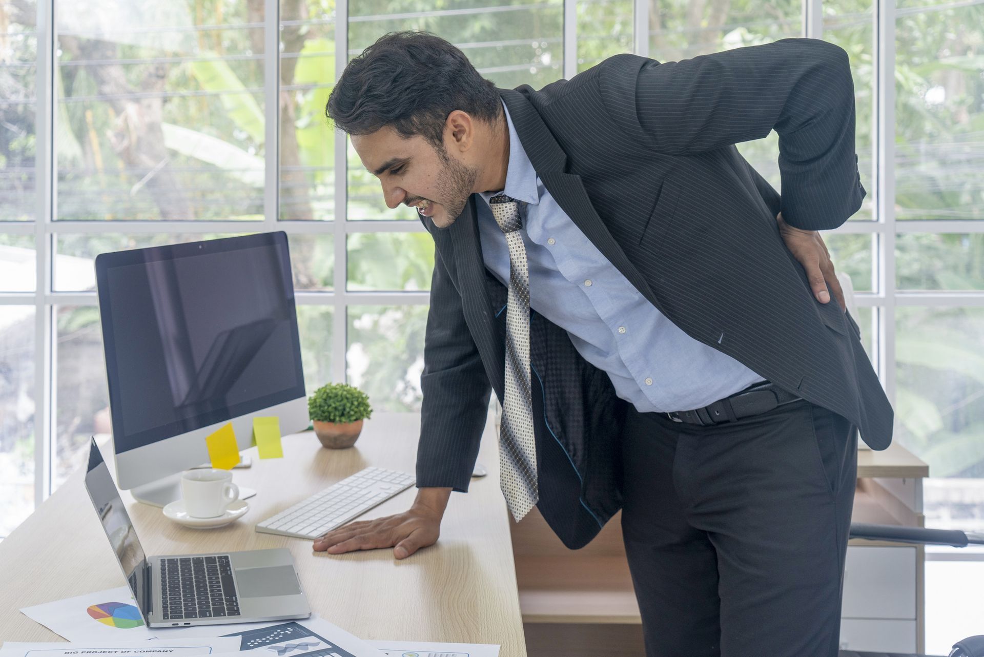 A man in a suit and tie is standing in front of a computer desk holding his back.