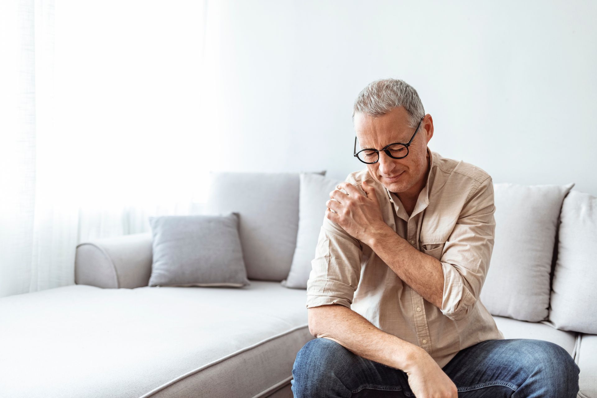 A man is sitting on a couch holding his shoulder in pain.