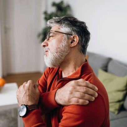 A man with a beard is sitting on a couch holding his shoulder in pain.