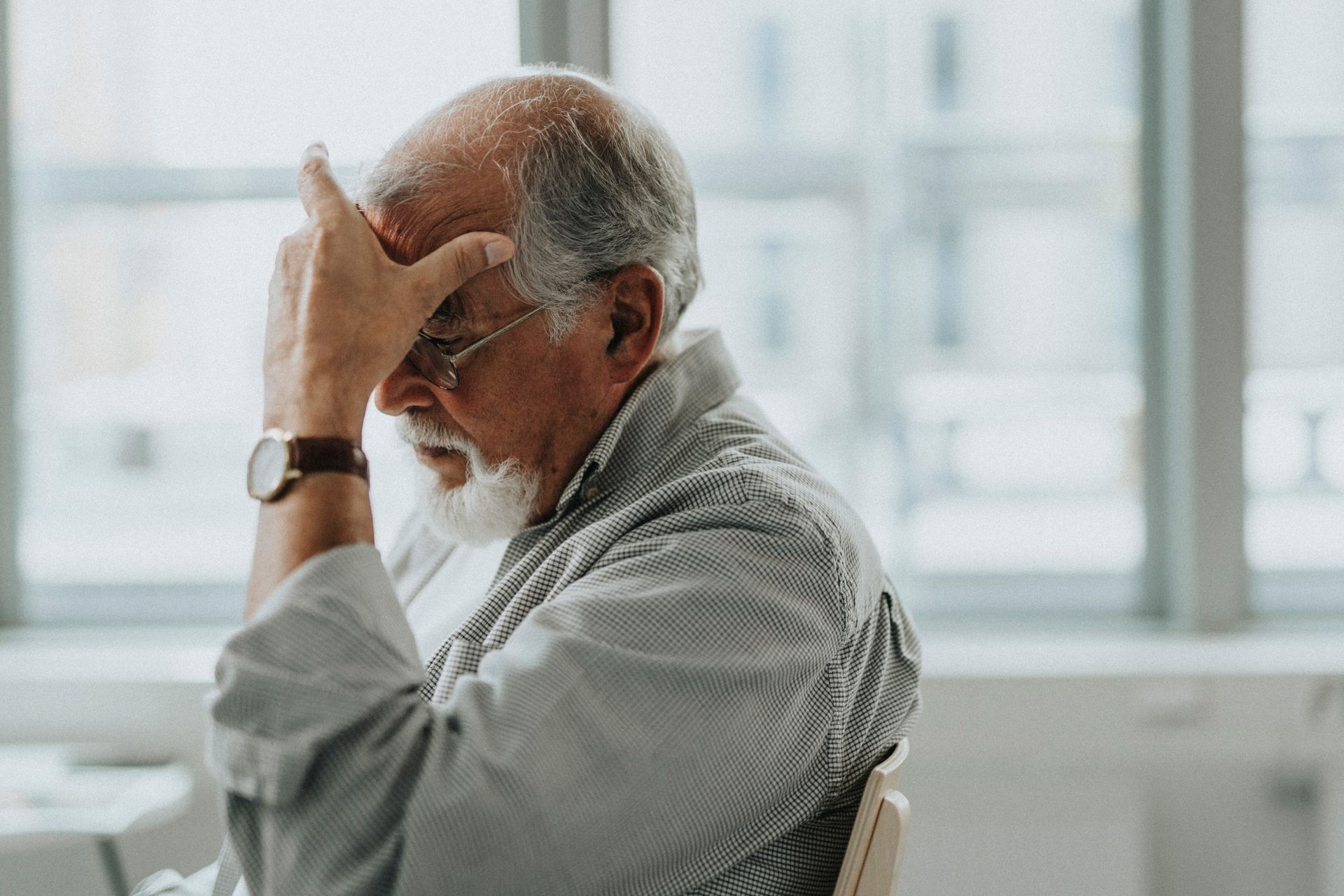 An elderly man is sitting in a chair with his hand on his head.