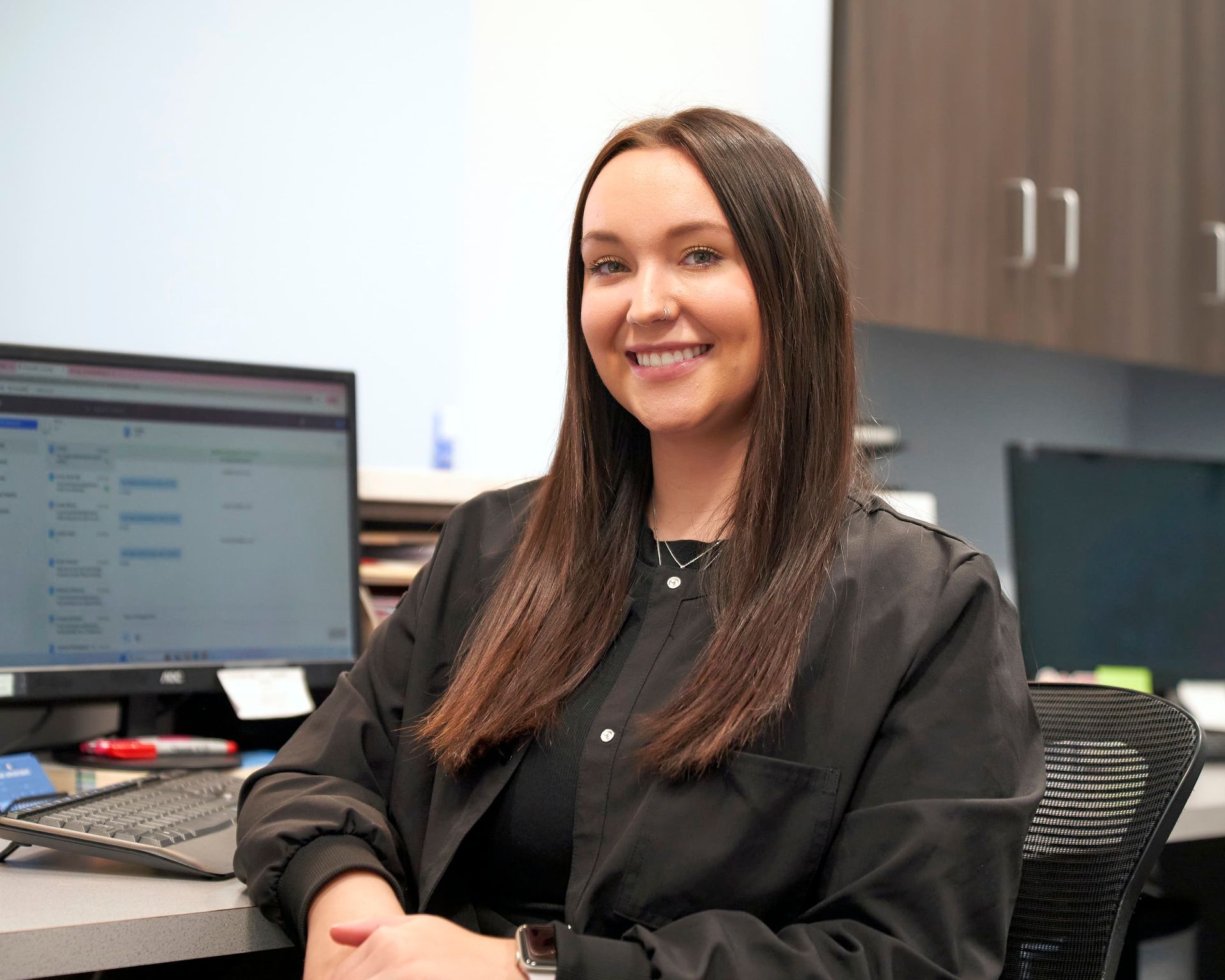 Woman with long brown hair smiling, sitting at a desk with a computer in an office setting.